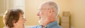 Elderly couple with glasses is looking at each other with joy with moving boxes in the far-right hand side