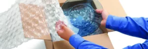 Man, in a blue shirt opening up a box filled with bull wrap and a clear plate